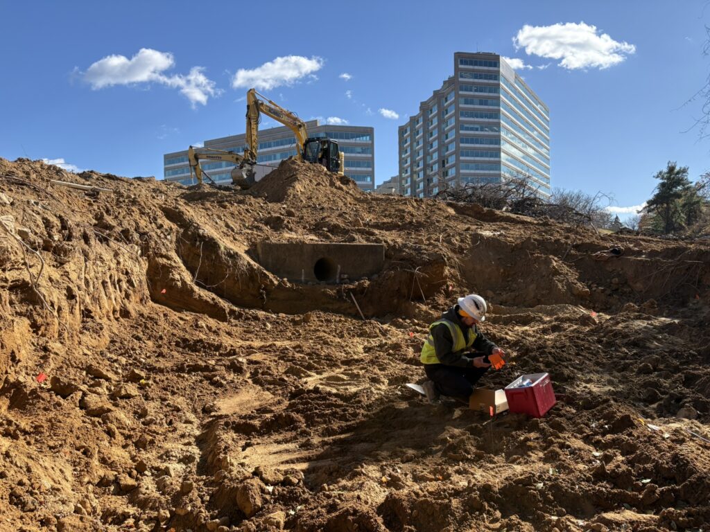 Construction site with a yellow excavator on a dirt mound and a worker in a hard hat nearby city buildings in the background