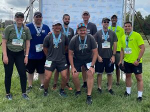 Group of runners wearing medals posing for a photo on grass with a race banner behind them.