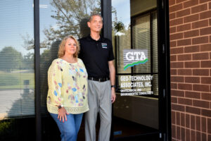 Two smiling adults stand outside a brick office building by a glass door with a GEO-TECHNOLOGY sign in the window.