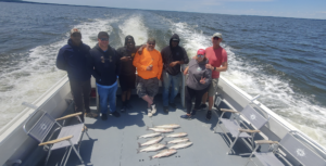 Group of people on a boat posing with a string of caught fish on the deck, wind in the water behind them.