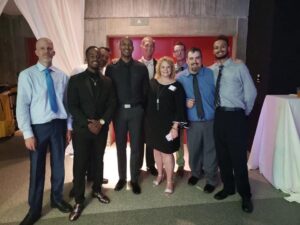 Eight adults in formal attire posing for a group photo at an indoor event with draped backdrop.