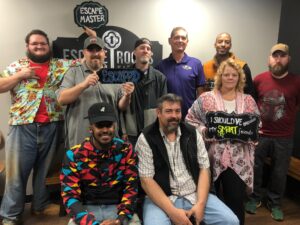 Group of eight adults posing in an escape room, with an 'Escaped' sign visible in the background and an Escape Master logo on the wall.