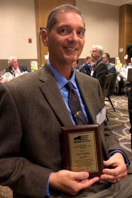 Smiling man in a brown blazer at a banquet, holding a Maryland Building Industry Association award plaque.