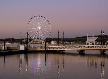 Colorful illuminated Ferris wheel at a harbor during sunset, its lights reflecting on the calm water.